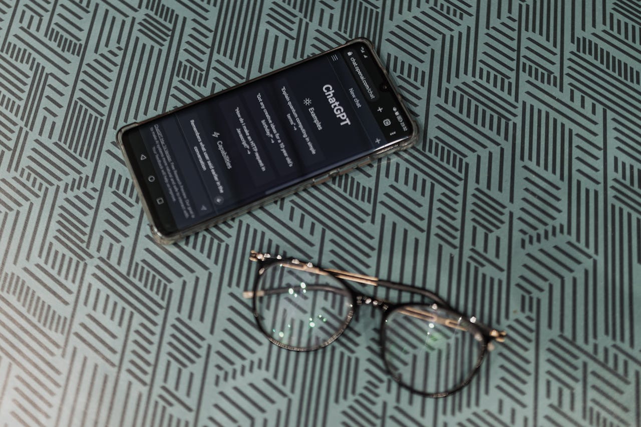 Close-up of smartphone displaying ChatGPT alongside eyeglasses on a patterned surface highlighting technology and style.